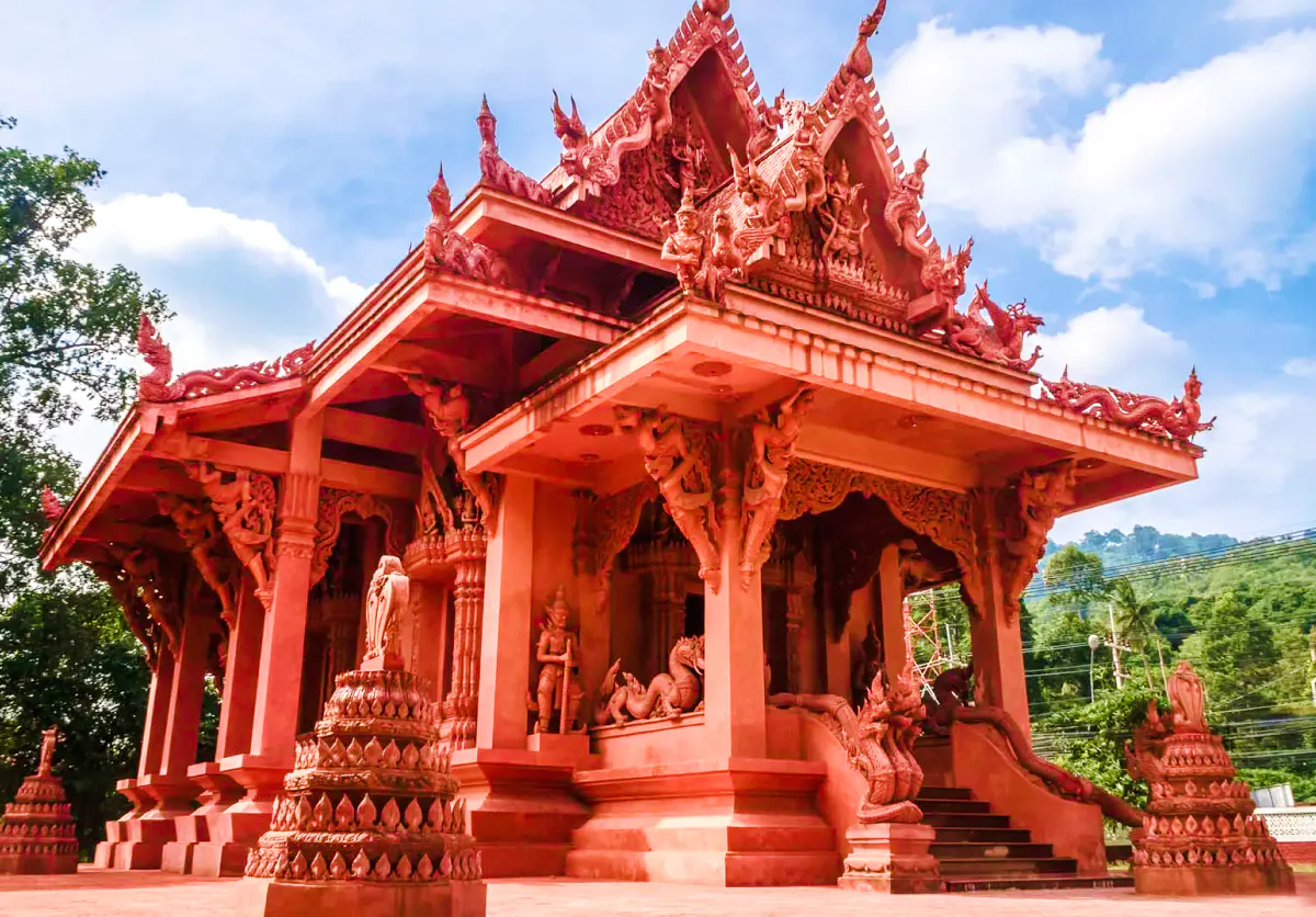 Red temple with intricate carvings in Koh Samui, surrounded by lush greenery and blue sky.