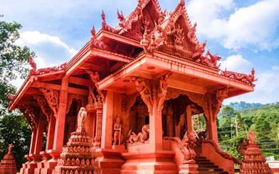Red temple with intricate carvings in Koh Samui, surrounded by lush greenery and blue sky.