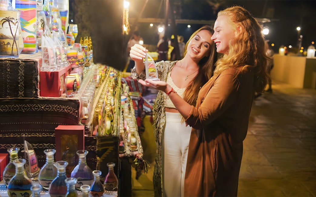 Women browsing colorful sand art bottles at a market during Royal dinner, Dubai.