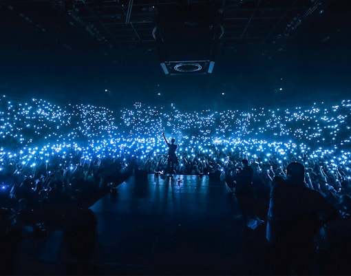 Performer on stage at a packed stadium concert with audience holding lights.