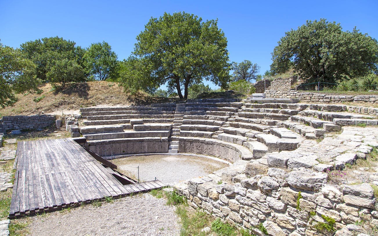 Ancient amphitheater ruins in Troy, Turkey, surrounded by trees under a clear sky.
