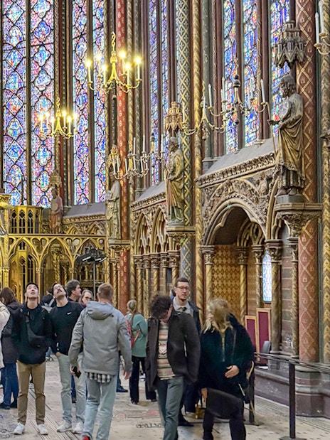 Visitors inside Sainte Chapelle, Paris, admiring stained glass windows and ornate Gothic architecture.