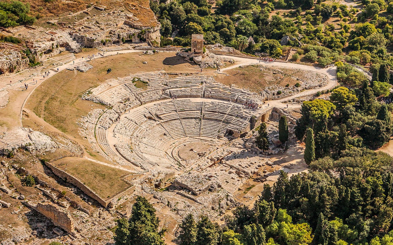 Aerial view of Syracuse's Greek Theater in Neapolis, Italy, surrounded by lush greenery.