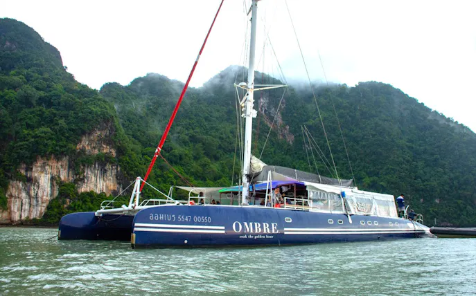 Catamaran Ombre Yacht sailing near lush cliffs in Phuket, Thailand.