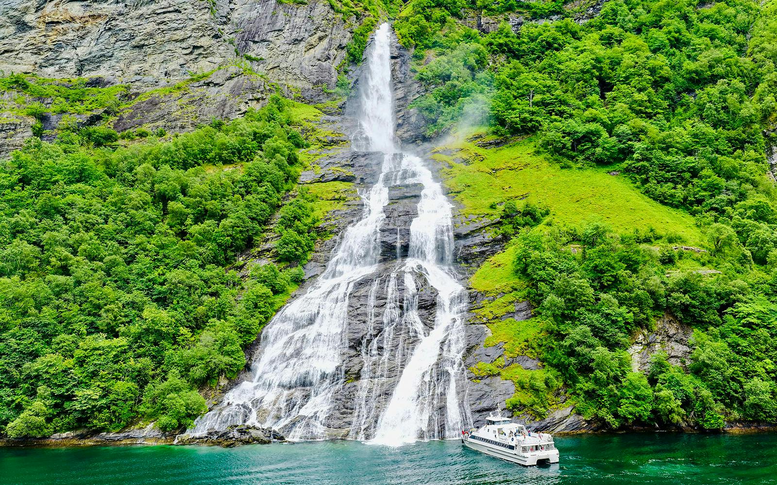 Boat near waterfall in Geiranger Fjord, Norway, surrounded by lush greenery.