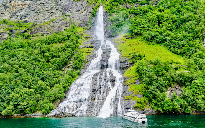 Boat near waterfall in Geiranger Fjord, Norway, surrounded by lush greenery.