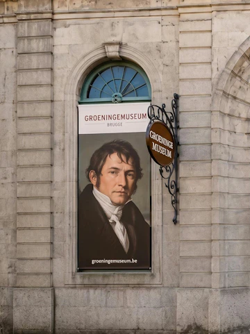 Banner of Groeninge Museum in Brugge with a portrait displayed on a stone building wall.