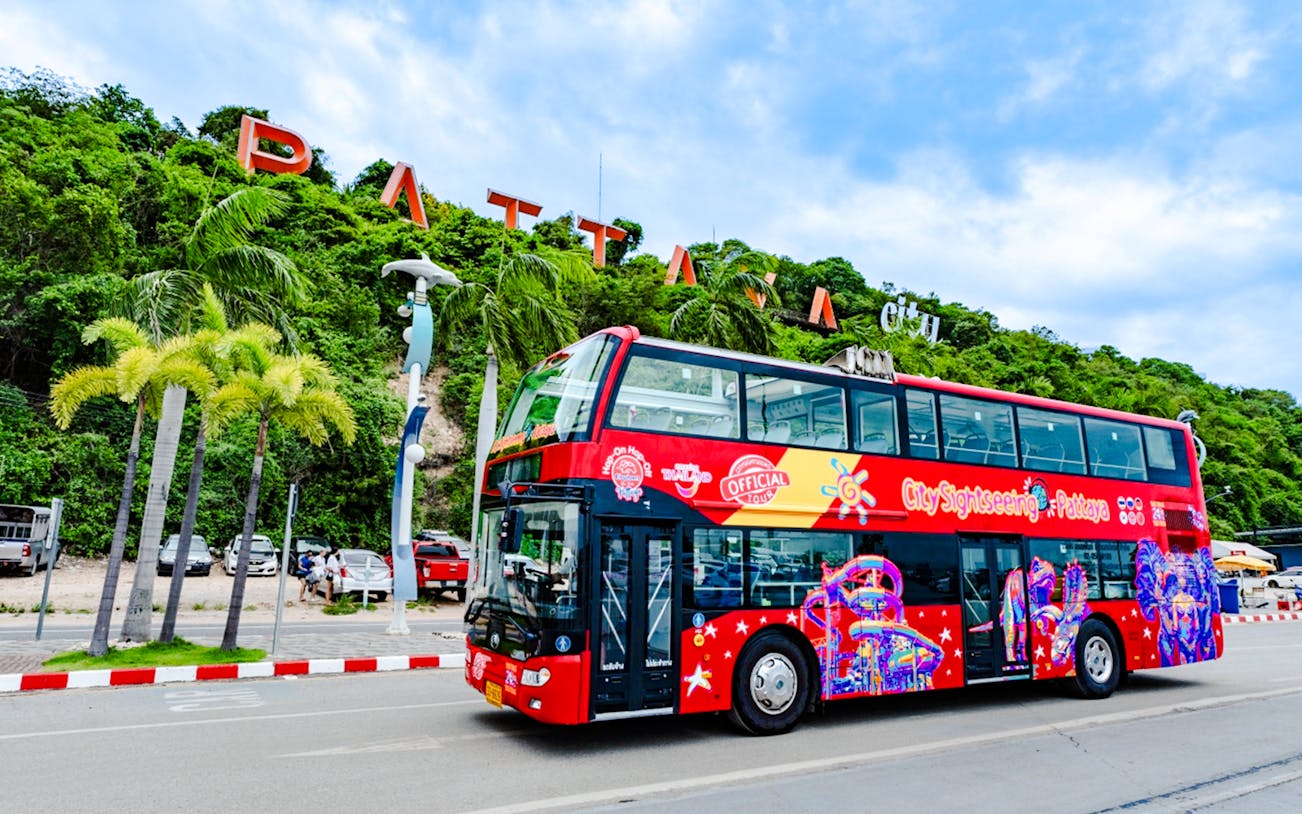 Pattaya hop on hop off bus passing by Pattaya sign with lush greenery.