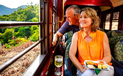 Couple enjoying scenic train tour with snacks and wine, lush landscape outside.