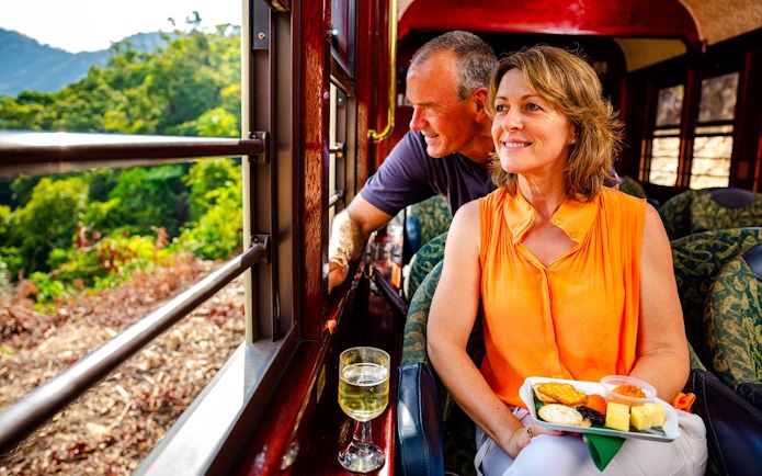 Couple enjoying scenic train tour with snacks and wine, lush landscape outside.