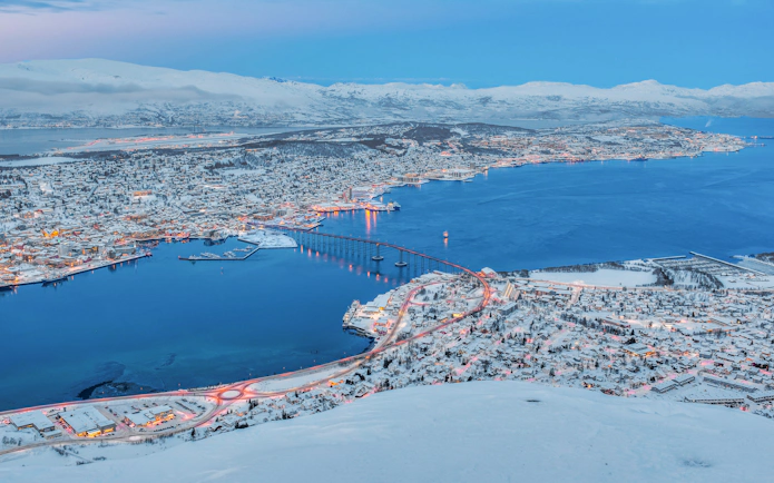 Aerial view of Tromso, Norway, with snow-covered landscape and bridge at sunset in winter.
