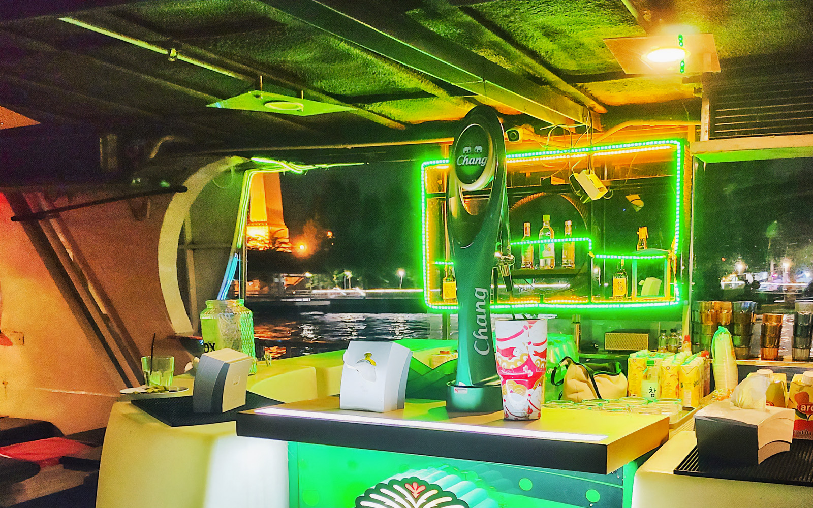 Bar area on Yodsiam sightseeing boat with drinks and illuminated backdrop, Bangkok river view.