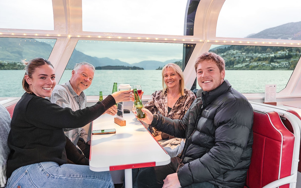 Passengers toast with drinks inside a cruise boat cabin, Queenstown scenery visible through the window.