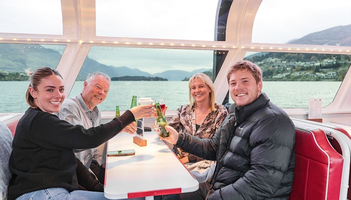Passengers toast with drinks inside a cruise boat cabin, Queenstown scenery visible through the window.