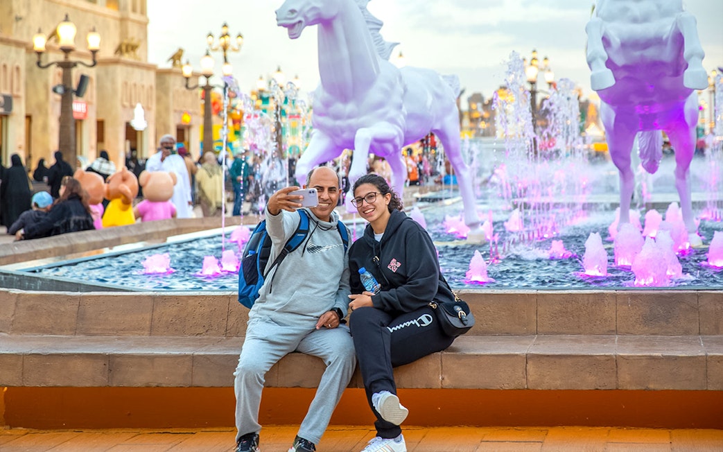 Visitors taking a selfie at Dubai Global Village with colorful fountain and horse statues.