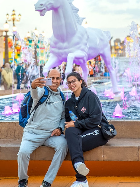 Visitors taking a selfie at Dubai Global Village with colorful fountain and horse statues.