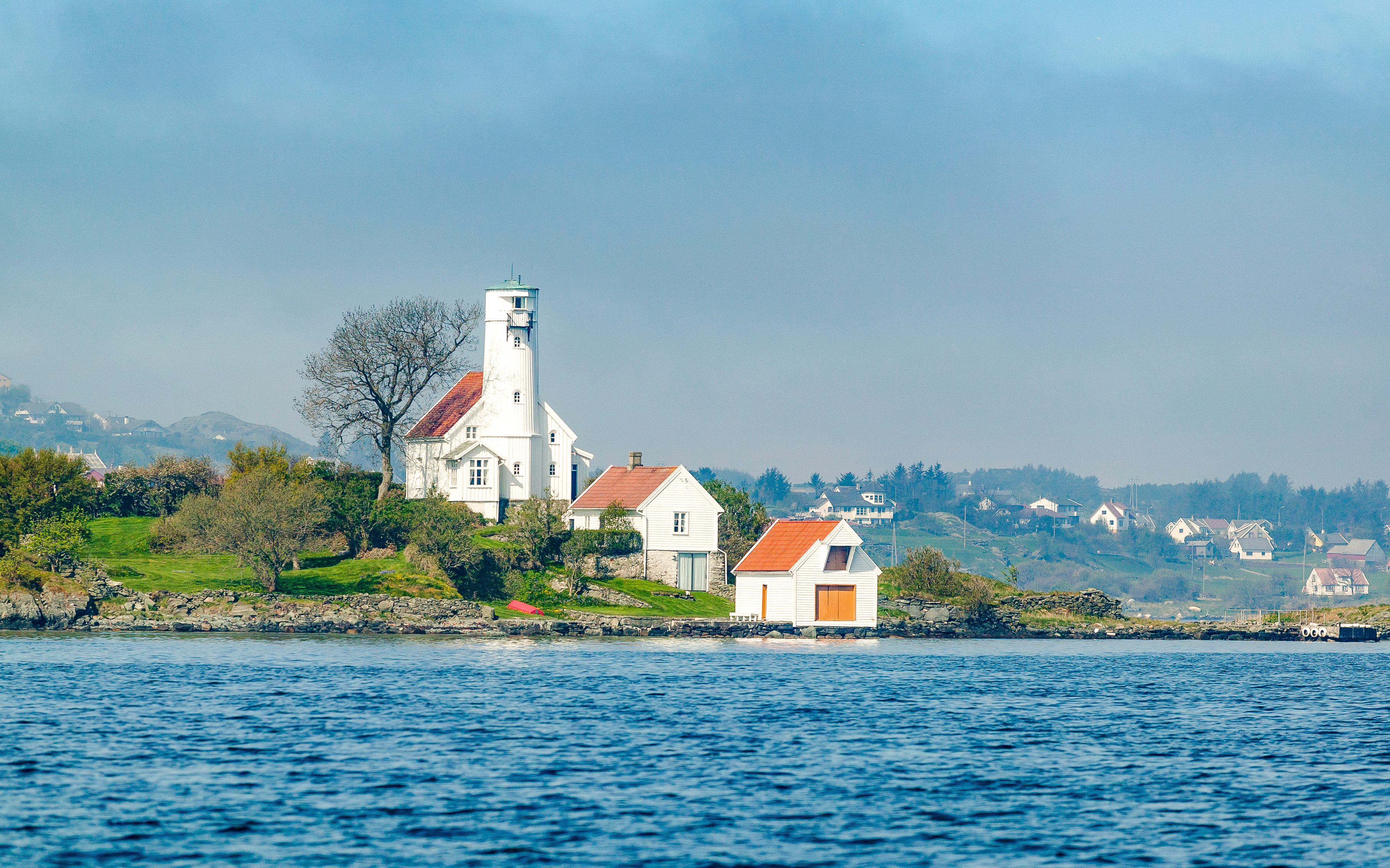 Haugesund lighthouse on a grassy coastline in Norway, with surrounding houses and calm water.
