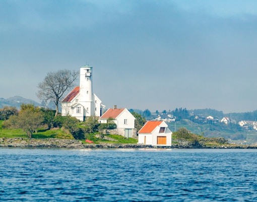 Haugesund lighthouse on a grassy coastline in Norway, with surrounding houses and calm water.
