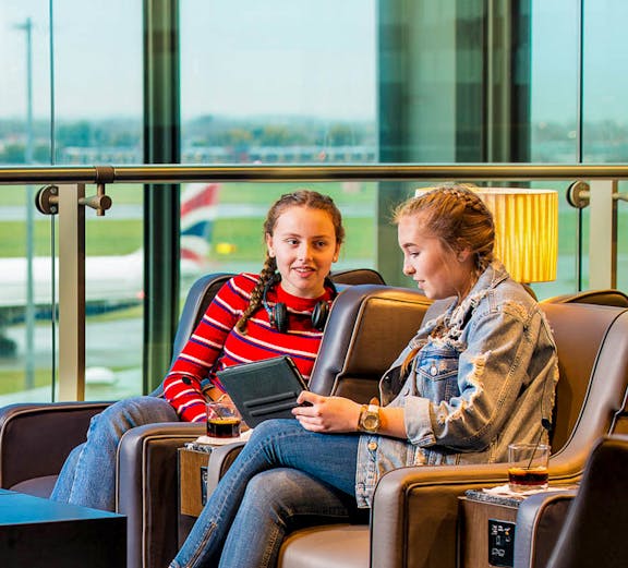 Two travelers relaxing in Plaza Premium Lounge Heathrow, seated with drinks and a tablet.