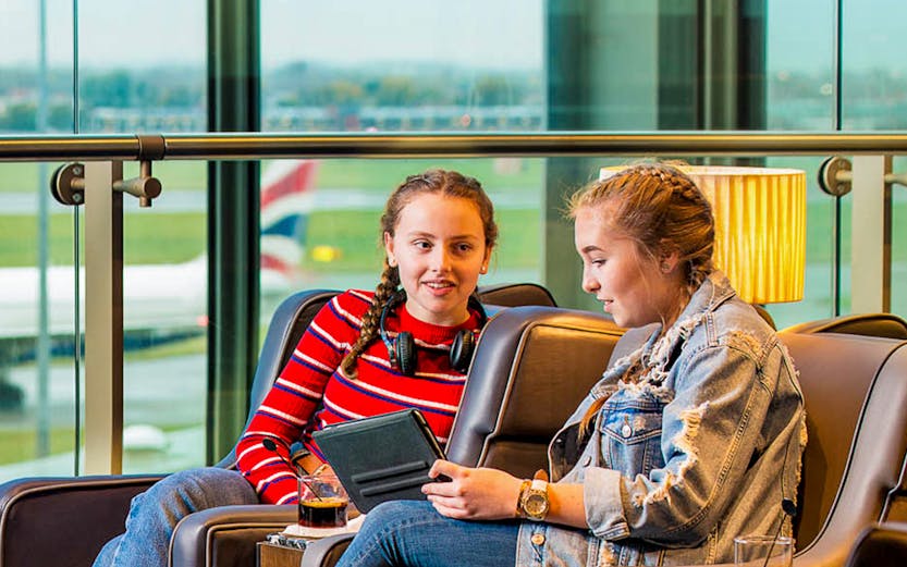 Two travelers relaxing in Plaza Premium Lounge Heathrow, seated with drinks and a tablet.