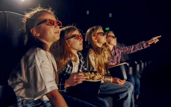 Kids wearing 3D glasses watching a movie at a theater, holding popcorn.