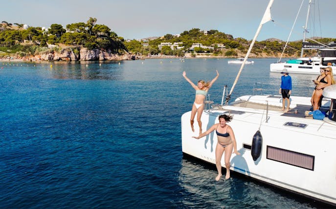 Two women jumping from catamaran LAGOO into blue water near a scenic coastline.