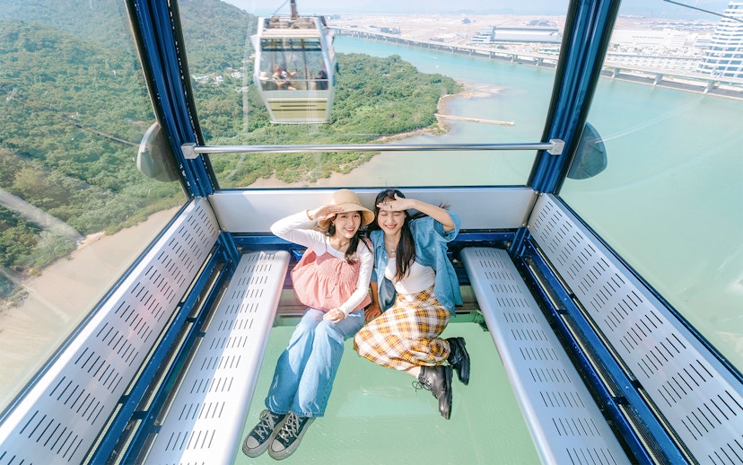 Two people sitting inside a crystal cabin cable car with a view of lush greenery and water below.