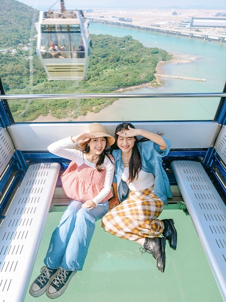 Two people sitting inside a crystal cabin cable car with a view of lush greenery and water below.