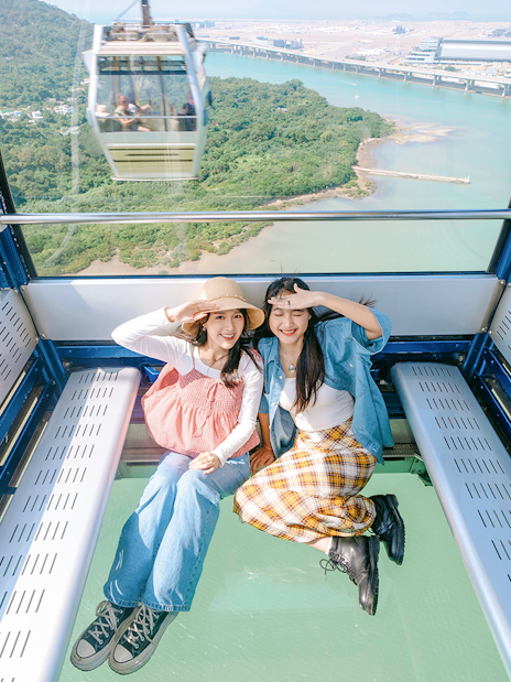 Two people sitting inside a crystal cabin cable car with a view of lush greenery and water below.