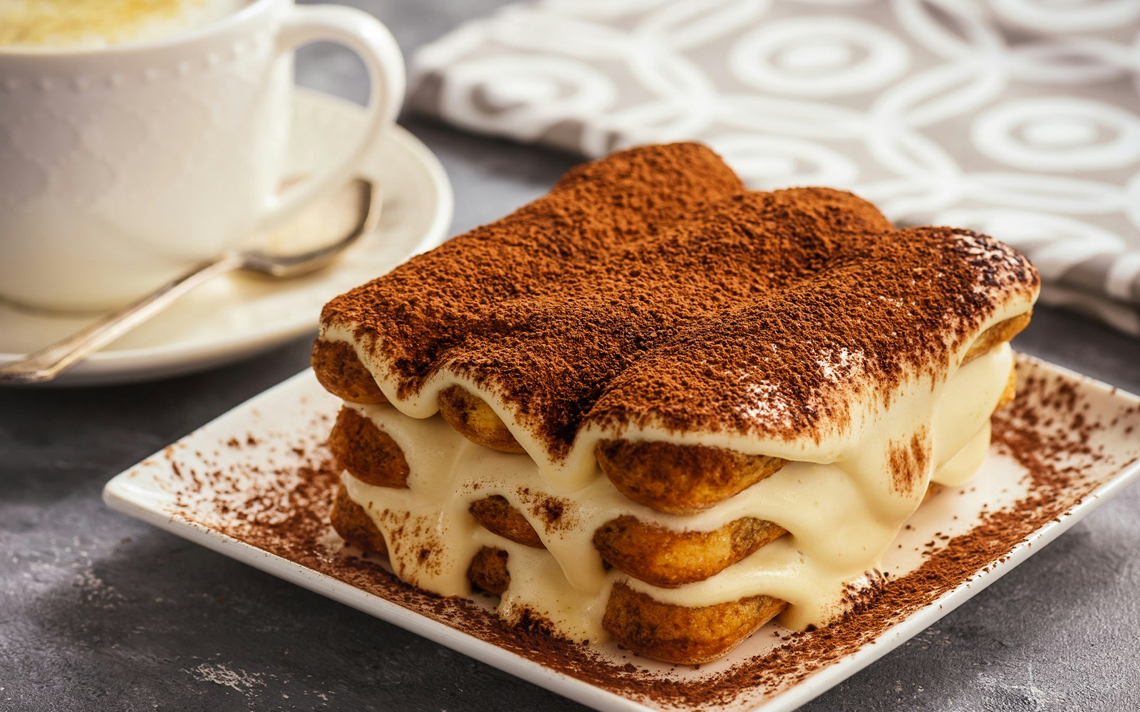 Tiramisu dessert with cocoa powder on a plate next to a cup of coffee.