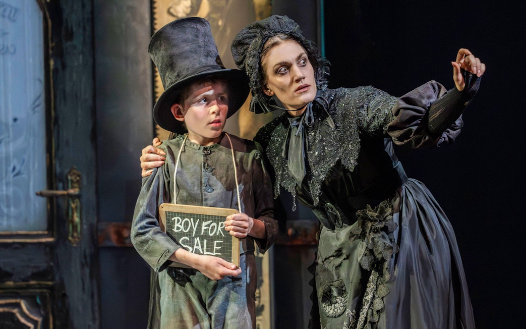 Child in top hat holding "Boy for Sale" sign with woman in period costume during Oliver production.