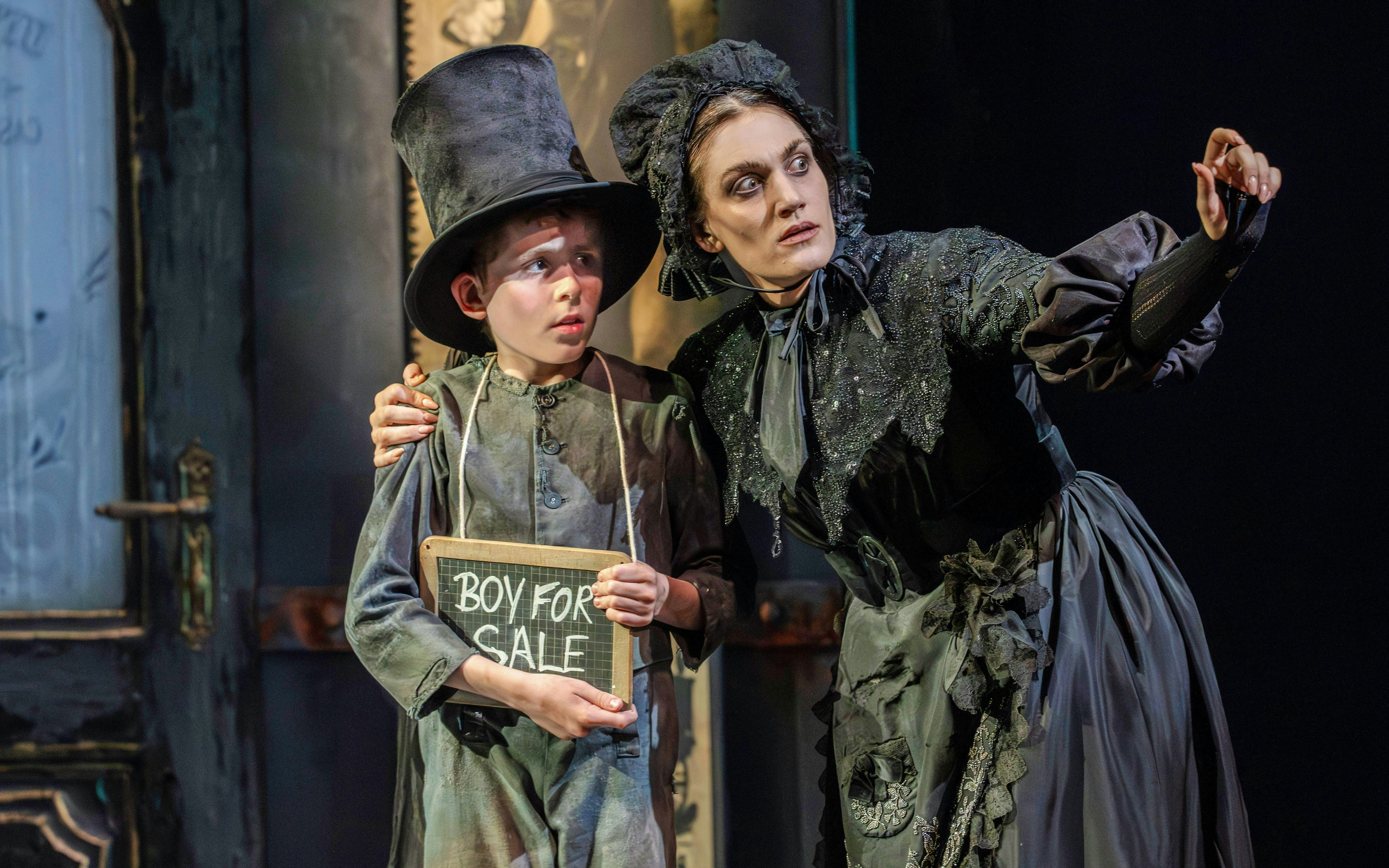 Child in top hat holding "Boy for Sale" sign with woman in period costume during Oliver production.