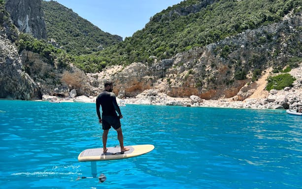 E-surfer gliding on turquoise waters in the Gulf of Alghero with rocky cliffs in the background.