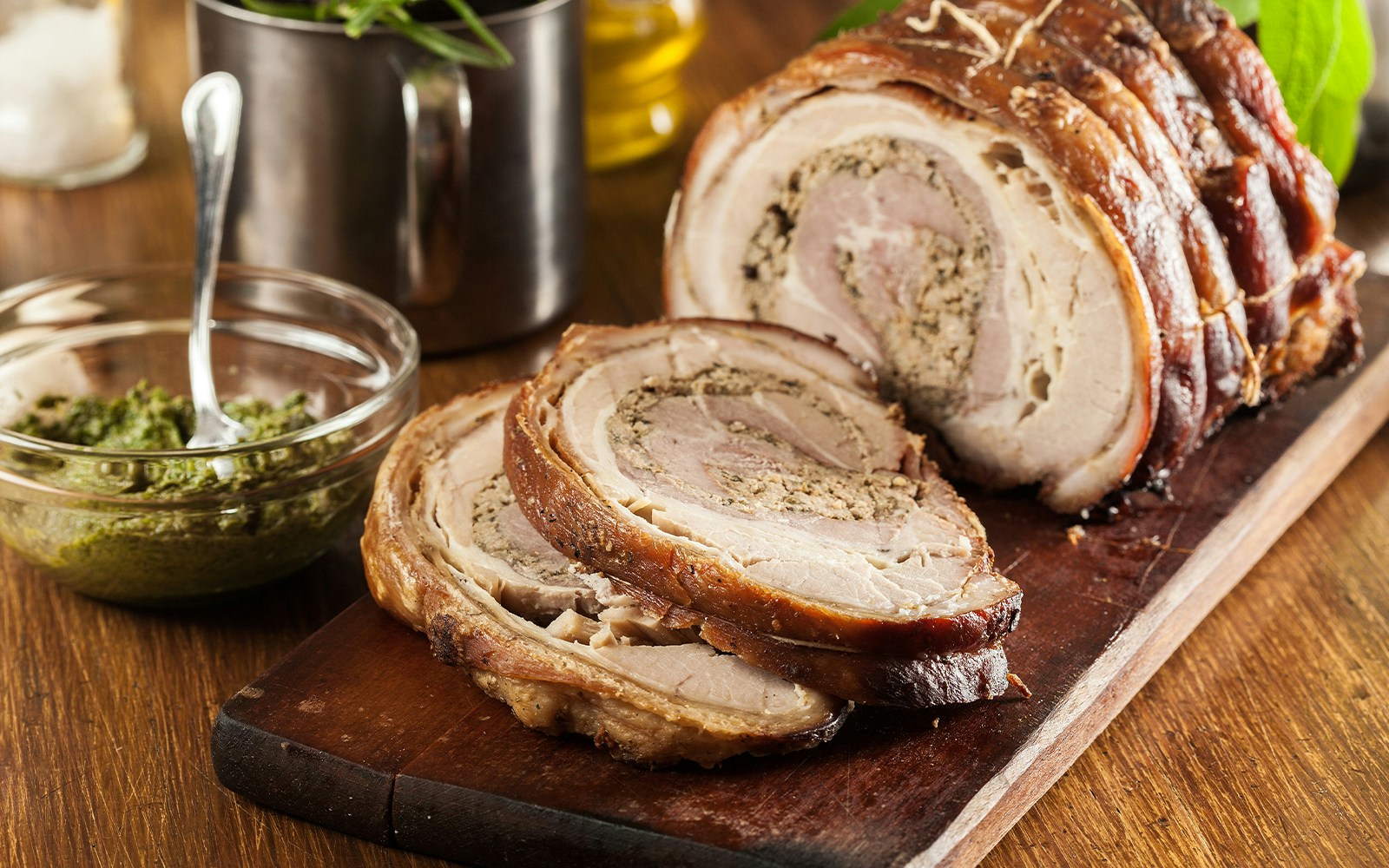 Sliced porchetta on a wooden board with a bowl of green sauce.