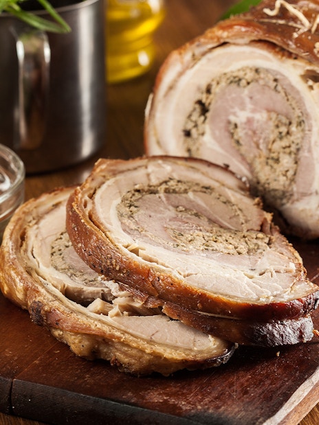 Sliced porchetta on a wooden board with a bowl of green sauce.