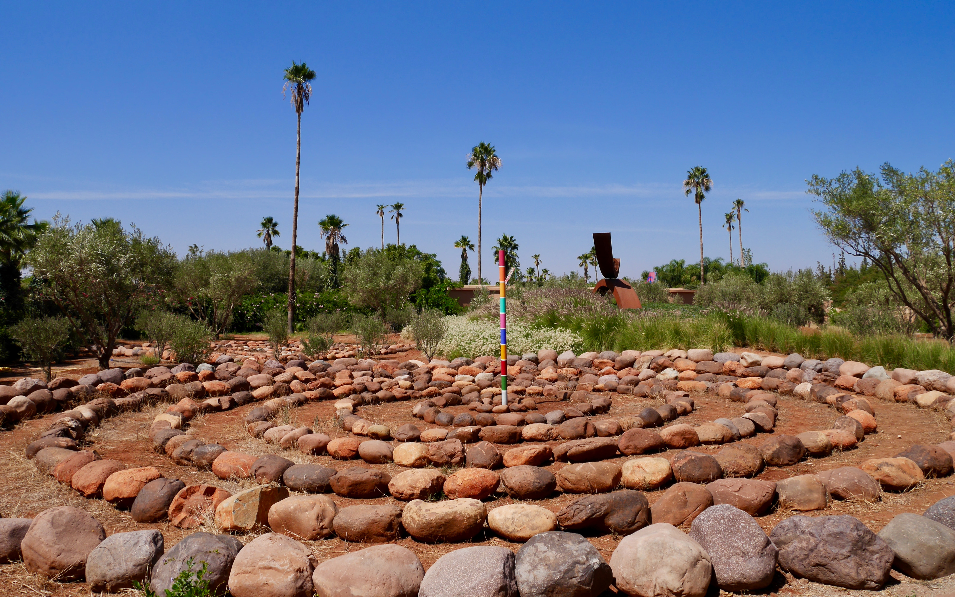 Stone labyrinth with colorful pole in ANIMA Garden, Marrakech, surrounded by palm trees and greenery.