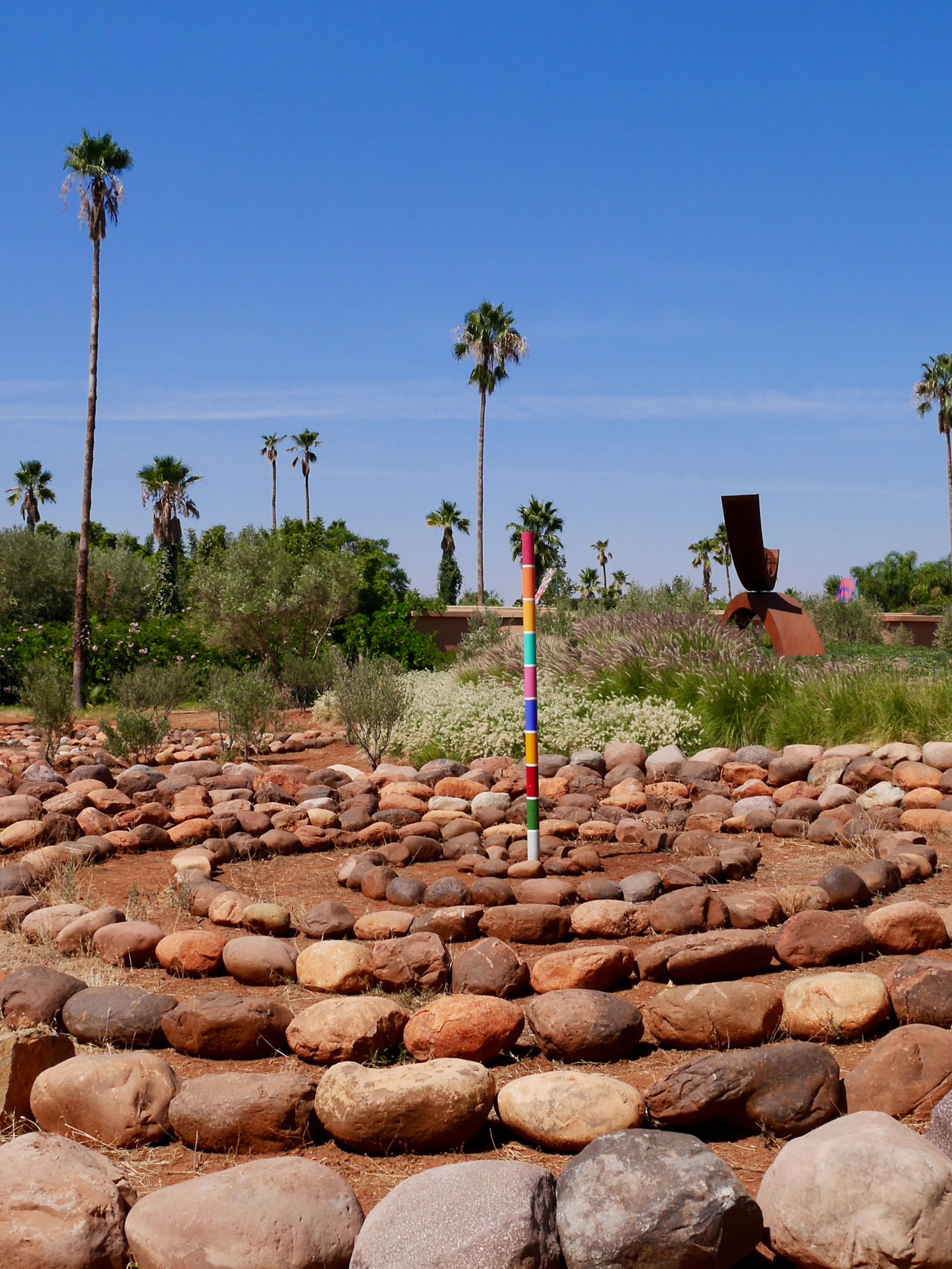 Stone labyrinth with colorful pole in ANIMA Garden, Marrakech, surrounded by palm trees and greenery.