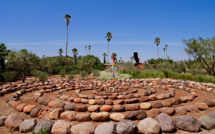 Stone labyrinth with colorful pole in ANIMA Garden, Marrakech, surrounded by palm trees and greenery.