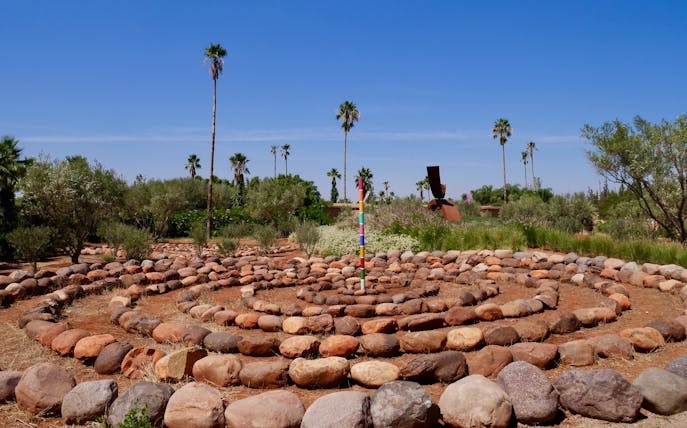 Stone labyrinth with colorful pole in ANIMA Garden, Marrakech, surrounded by palm trees and greenery.