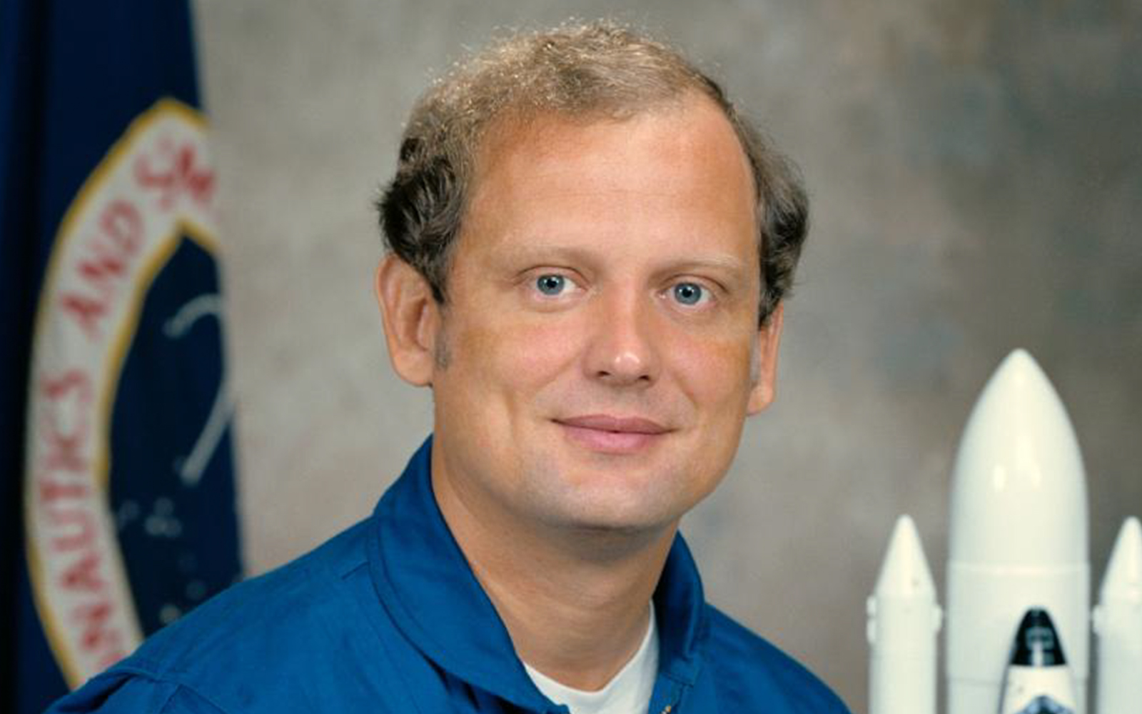 Norm Thagard in astronaut suit with NASA patch and American flag backdrop.