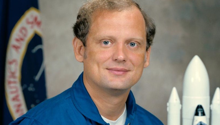 Norm Thagard in astronaut suit with NASA patch and American flag backdrop.