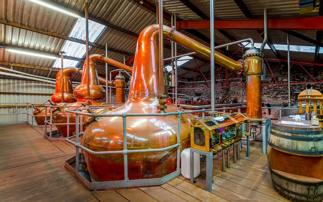 Copper stills inside a whiskey distillery with wooden barrels and equipment.