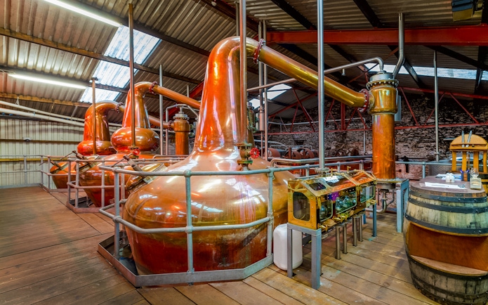 Copper stills inside a whiskey distillery with wooden barrels and equipment.