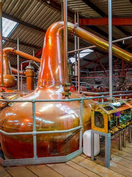 Copper stills inside a whiskey distillery with wooden barrels and equipment.