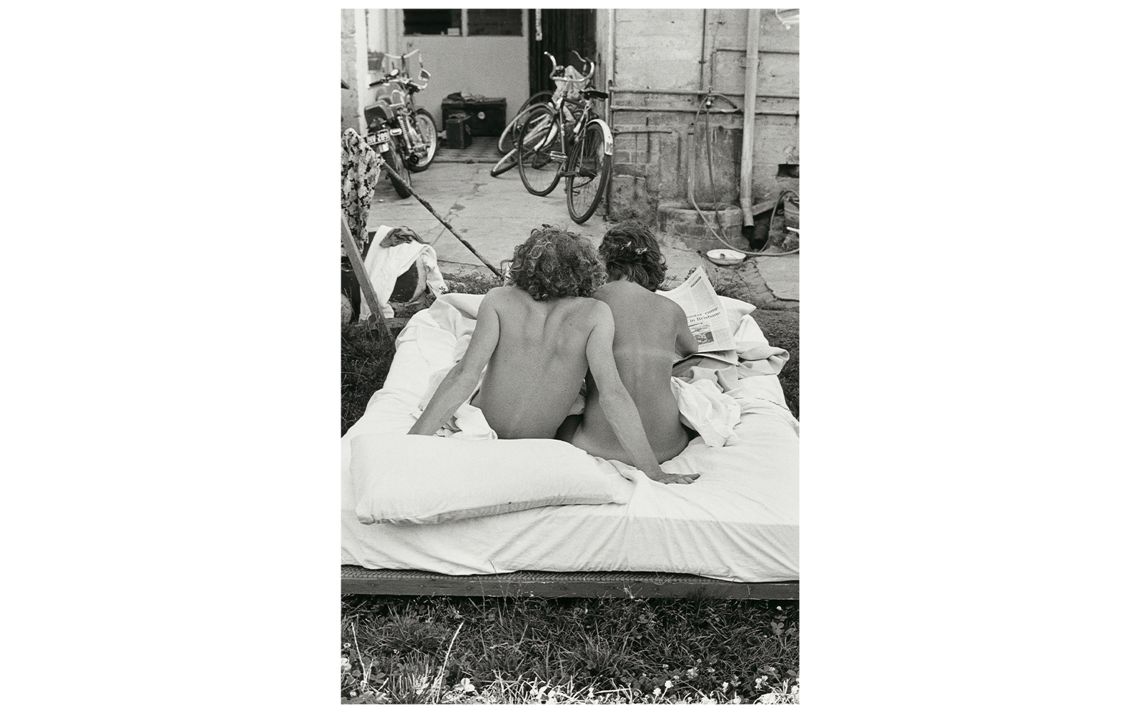 Two people sitting on an outdoor mattress, reading a newspaper, with bicycles in the background.