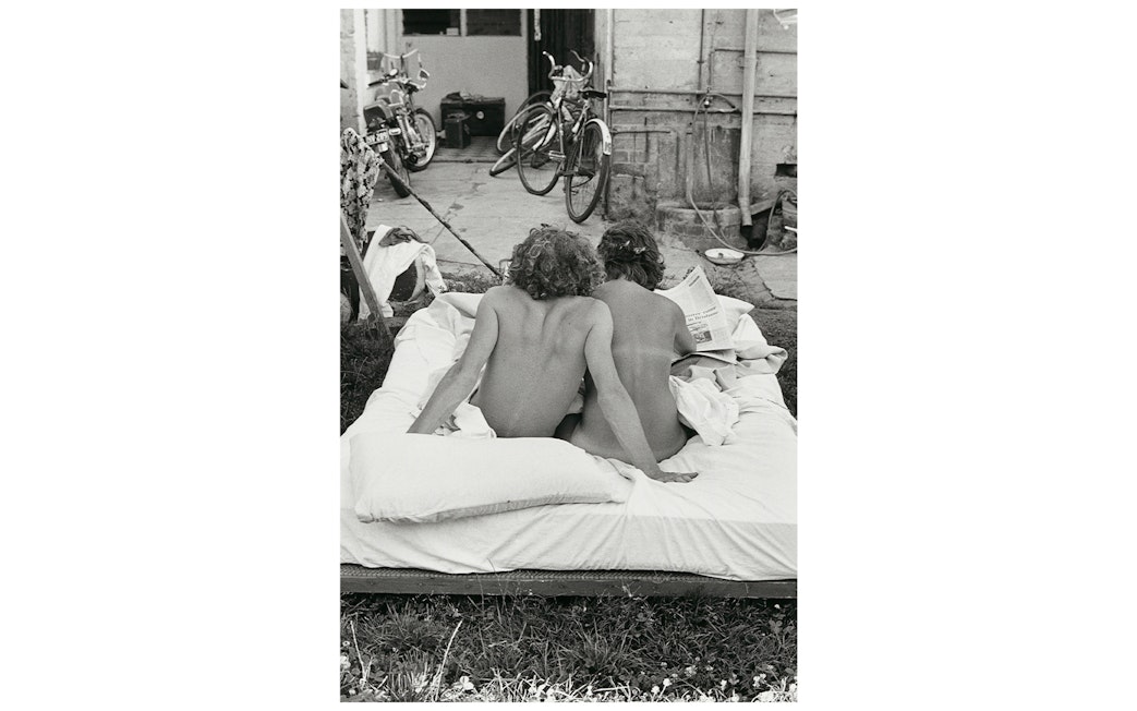 Two people sitting on an outdoor mattress, reading a newspaper, with bicycles in the background.