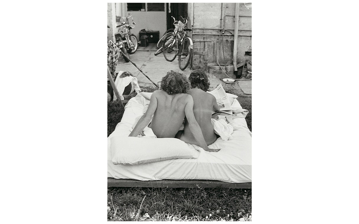 Two people sitting on an outdoor mattress, reading a newspaper, with bicycles in the background.