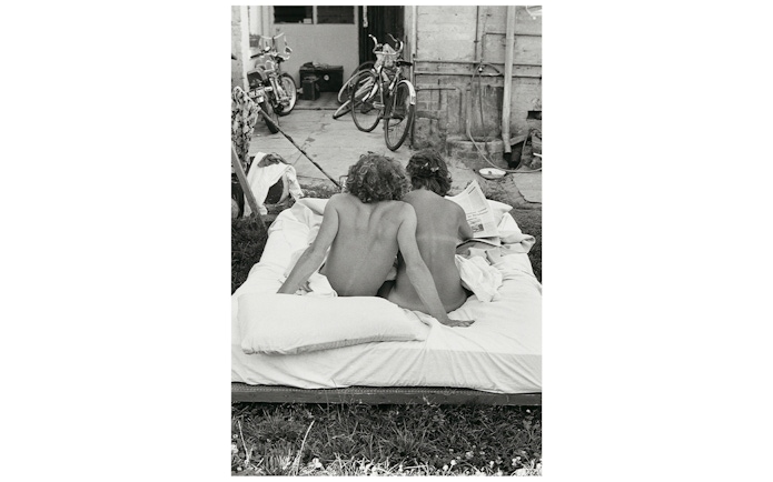 Two people sitting on an outdoor mattress, reading a newspaper, with bicycles in the background.