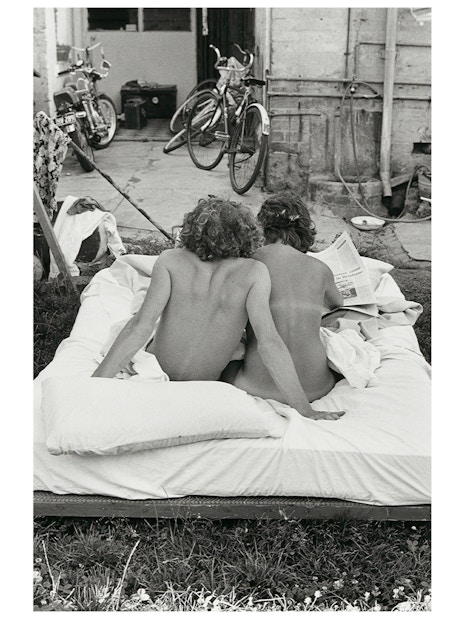 Two people sitting on an outdoor mattress, reading a newspaper, with bicycles in the background.