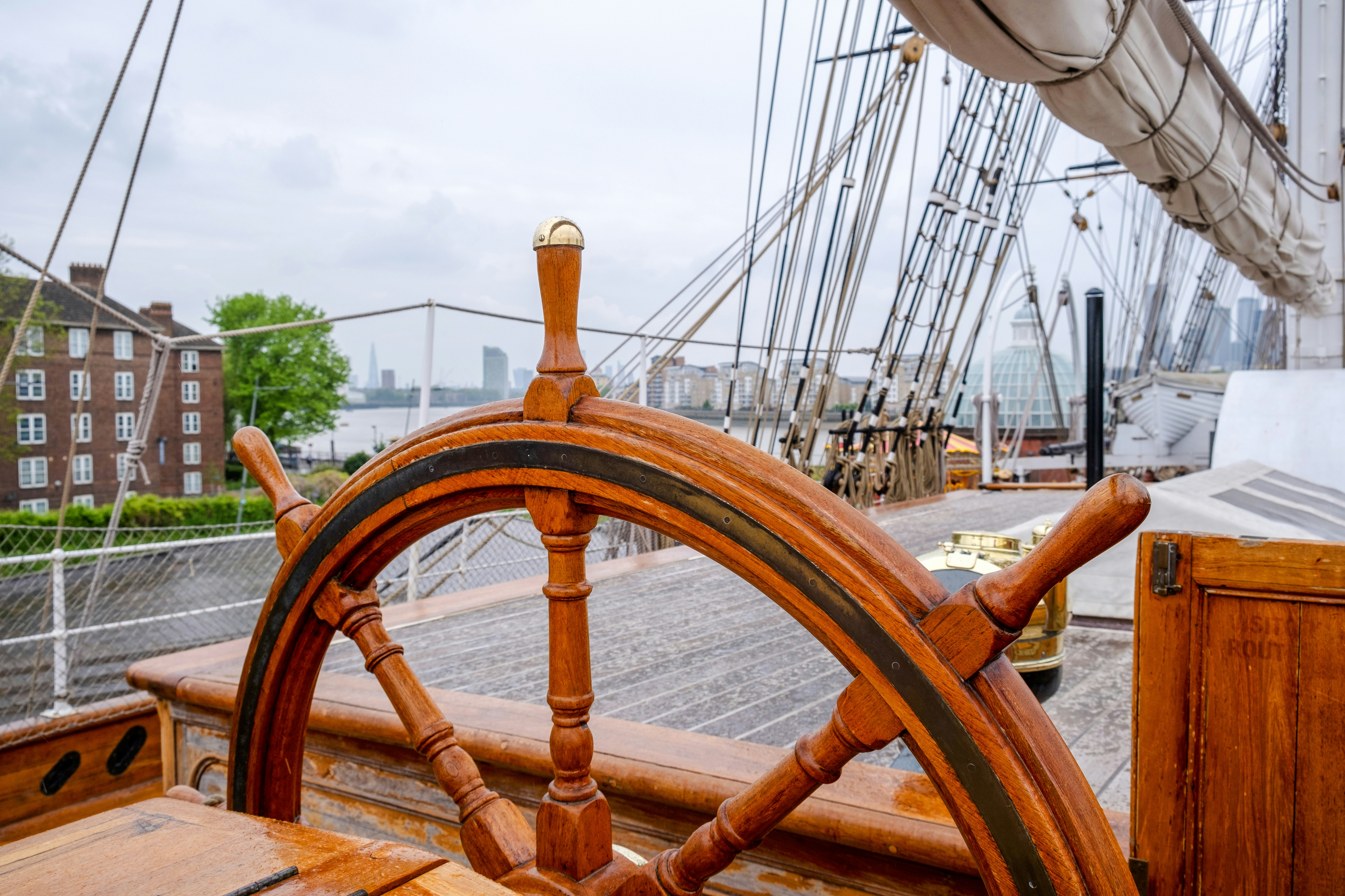 Ship wheel on a historic sailing vessel docked in a city harbor.